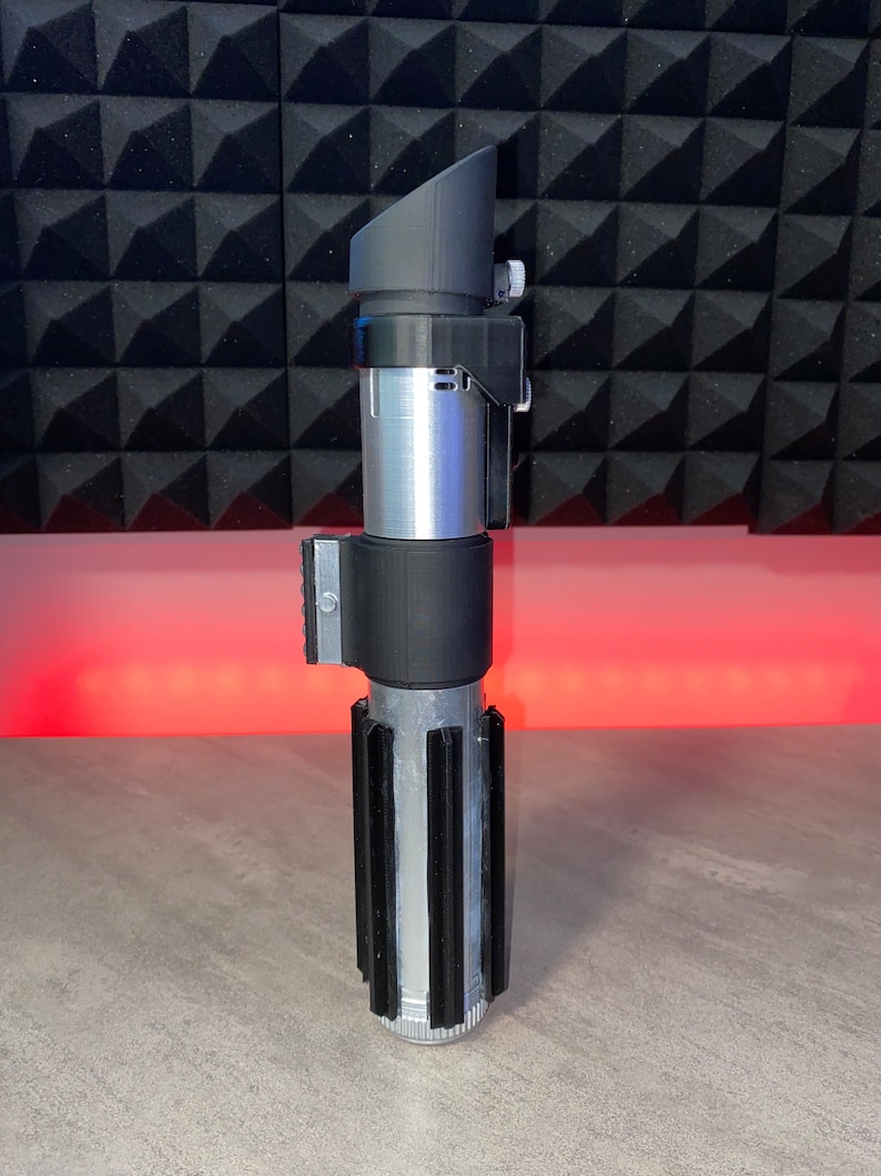 May include: A silver and black lightsaber prop with a rectangular viewing scope. The hilt has a ribbed design with black accents. The prop is made of plastic and metal and sits on a gray surface.