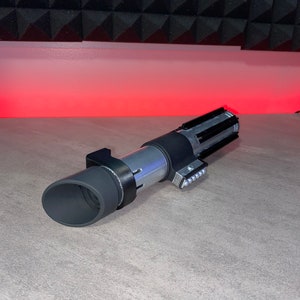 May include: A silver and black lightsaber prop with a cylindrical body and detailed handle. The prop features a black grip, a silver midsection, and a black emitter. The lightsaber is lying on a gray surface.