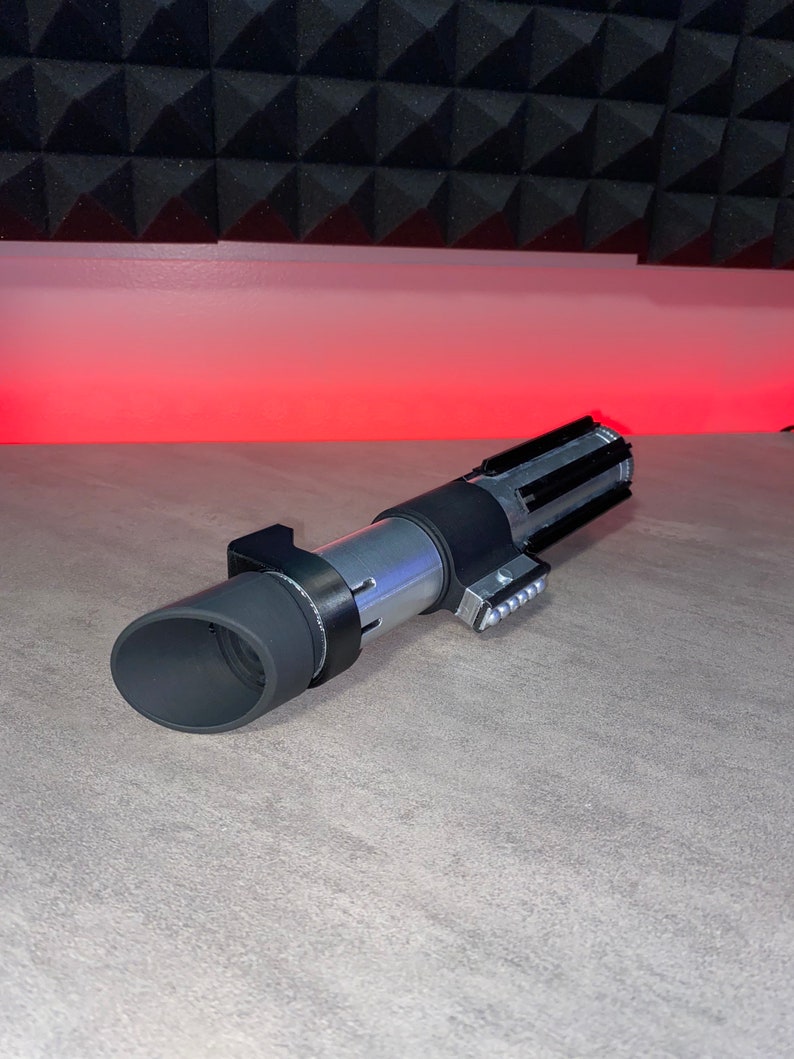 May include: A silver and black lightsaber prop with a cylindrical body and detailed handle. The prop features a black grip, a silver midsection, and a black emitter. The lightsaber is lying on a gray surface.
