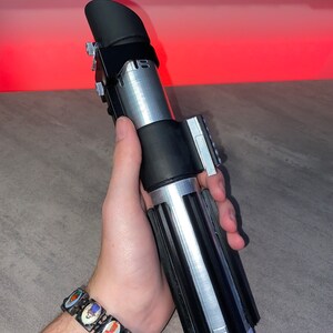 May include: A silver and black lightsaber prop with a black emitter and grip. The hilt has a ribbed design and silver accents. The lightsaber is held in a hand, with a colorful bracelet visible on the wrist.