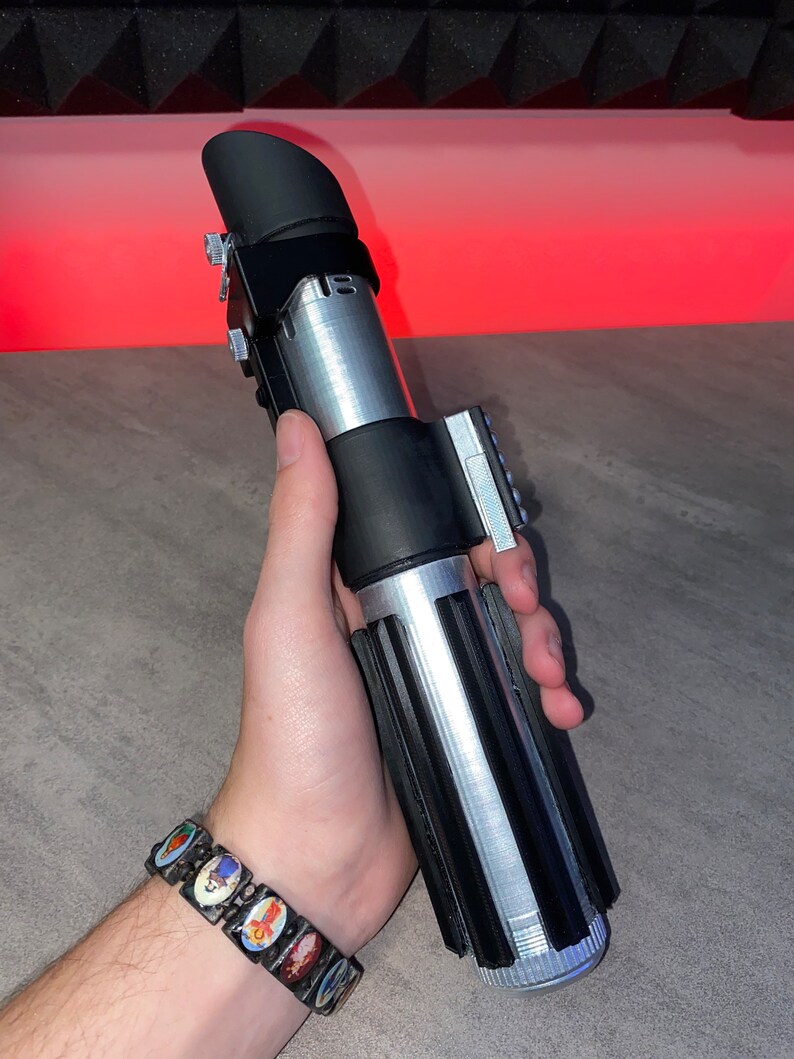 May include: A silver and black lightsaber prop with a black emitter and grip. The hilt has a ribbed design and silver accents. The lightsaber is held in a hand, with a colorful bracelet visible on the wrist.