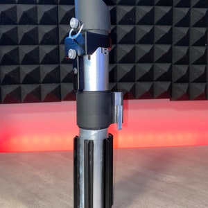 May include: A silver and black lightsaber prop with a ribbed handle and a black emitter. The prop has a cylindrical design with a black top and a silver body. The lightsaber is a replica of a popular science fiction weapon.