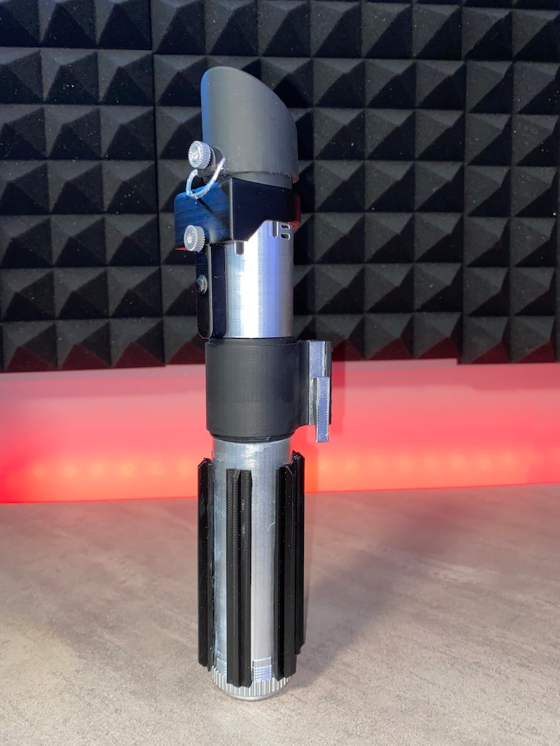 May include: A silver and black lightsaber prop with a ribbed handle and a black emitter. The prop has a cylindrical design with a black top and a silver body. The lightsaber is a replica of a popular science fiction weapon.