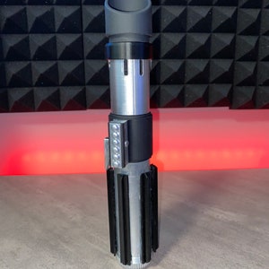 May include: A silver and black lightsaber prop with a cylindrical design. The hilt features a series of black fins and a silver body. The top has a gray cylindrical emitter. A row of silver lights is on the side.