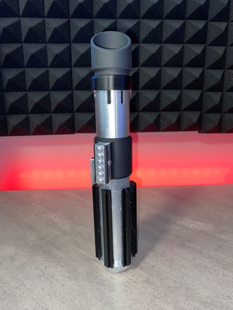 May include: A silver and black lightsaber prop with a cylindrical design. The hilt features a series of black fins and a silver body. The top has a gray cylindrical emitter. A row of silver lights is on the side.