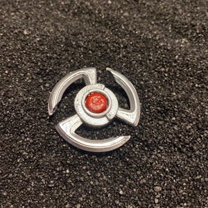 May include: A silver-colored metal toy with three arms and a red center. The toy is on a black background.