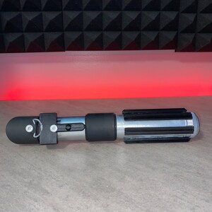 May include: A silver and black lightsaber prop with a black handle and emitter. The lightsaber has a silver blade section with black details. The prop is lying on a gray surface with a red light in the background.