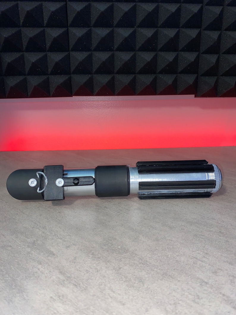 May include: A silver and black lightsaber prop with a black handle and emitter. The lightsaber has a silver blade section with black details. The prop is lying on a gray surface with a red light in the background.