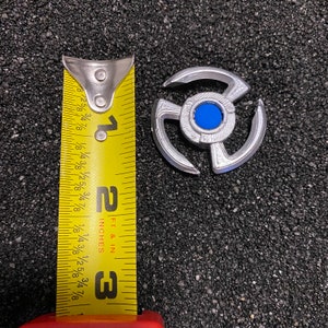 May include: A silver-colored metal Beyblade with a blue center and three arms. The Beyblade is about 1 inch in diameter and is next to a measuring tape.
