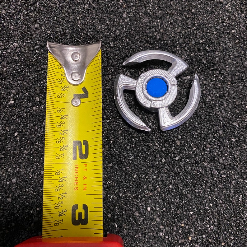 May include: A silver-colored metal Beyblade with a blue center and three arms. The Beyblade is about 1 inch in diameter and is next to a measuring tape.
