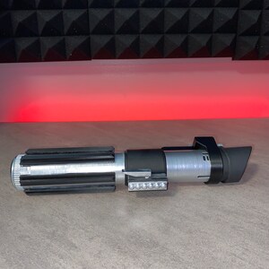 May include: A silver and black lightsaber prop with a detailed design. The hilt features a ribbed grip, a series of small lights, and a black emitter shroud. The prop is set against a gray surface with a red glow in the background.