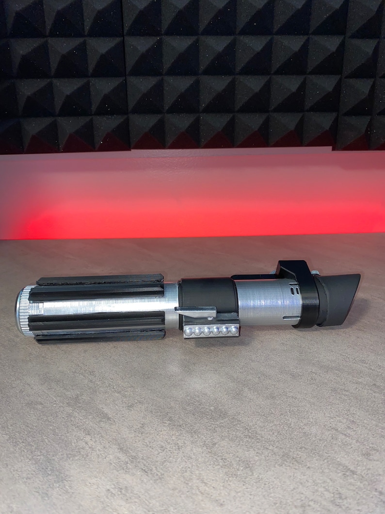 May include: A silver and black lightsaber prop with a detailed design. The hilt features a ribbed grip, a series of small lights, and a black emitter shroud. The prop is set against a gray surface with a red glow in the background.