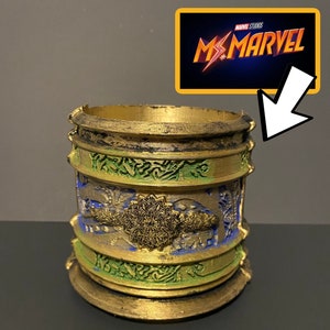 May include: A gold-coloured cylindrical container with intricate green and blue designs. The container has a textured surface and appears to be made of metal. The text "MARVEL STUDIOS MS. MARVEL" is visible in the background.
