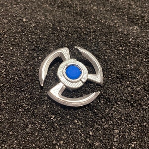 May include: A silver-colored metal spinning top with a blue center. The top has three arms that extend out from the center.