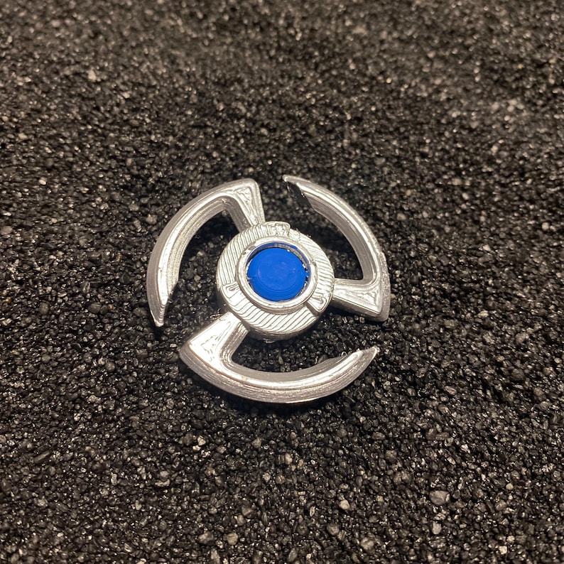 May include: A silver-colored metal spinning top with a blue center. The top has three arms that extend out from the center.
