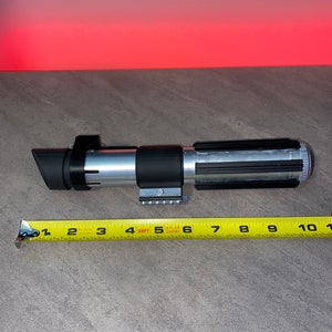 May include: A silver and black lightsaber prop, approximately 10 inches long, with a black emitter and a detailed handle. A yellow measuring tape is placed next to the prop.
