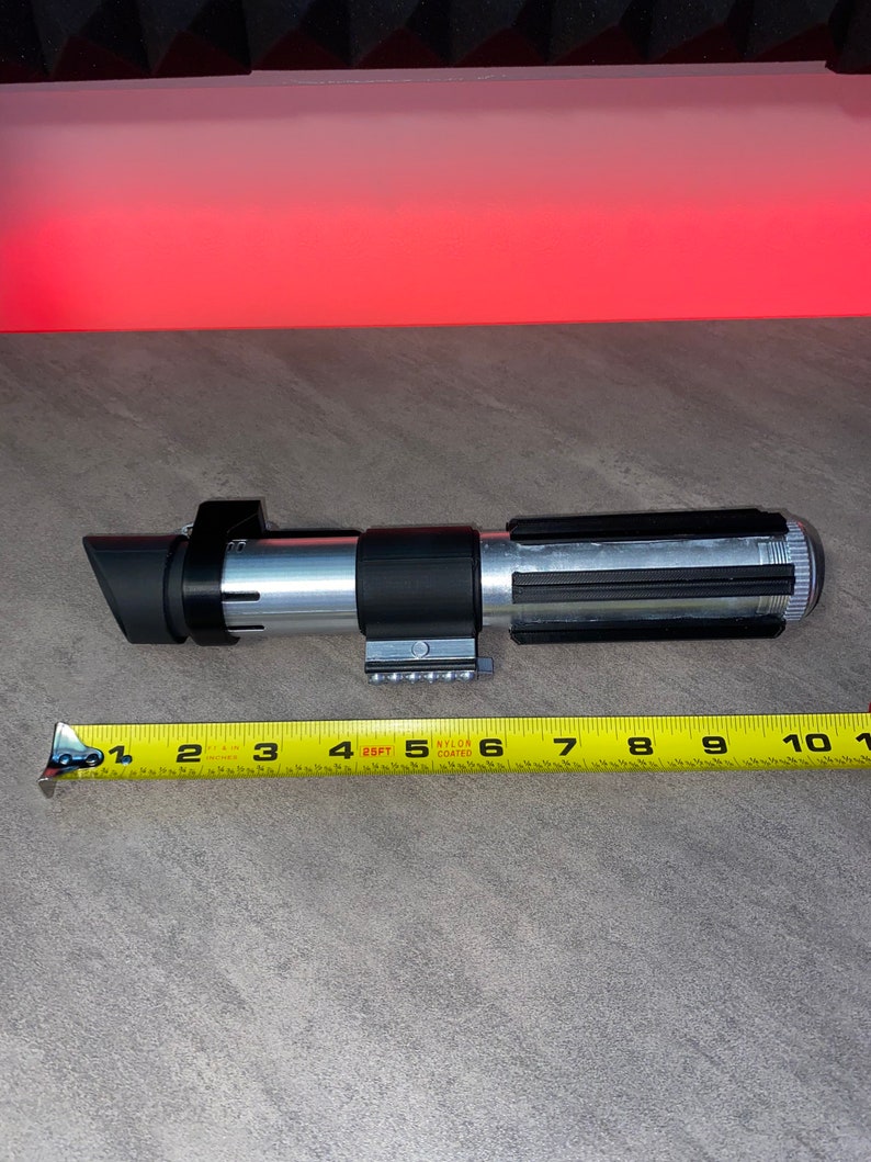 May include: A silver and black lightsaber prop, approximately 10 inches long, with a black emitter and a detailed handle. A yellow measuring tape is placed next to the prop.