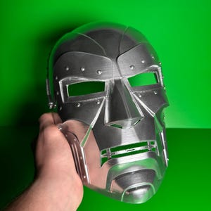 May include: A metallic silver-colored full-face mask featuring a rigid, angular design with prominent brow ridges, rectangular eye cutouts, and a horizontal mouth slit. The surface displays visible horizontal print lines and small circular rivet details.