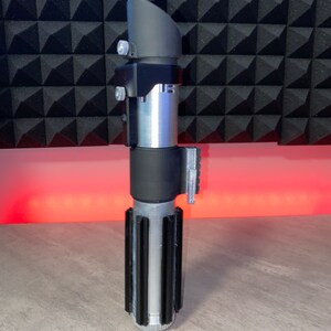 May include: A silver and black lightsaber prop with a cylindrical body and a black handle. The prop features a black emitter and a silver blade chamber. The lightsaber is a replica of a popular science fiction weapon.