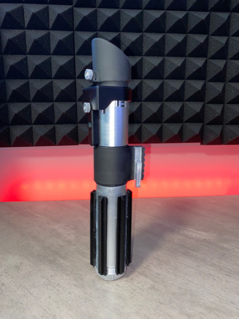 May include: A silver and black lightsaber prop with a cylindrical body and a black handle. The prop features a black emitter and a silver blade chamber. The lightsaber is a replica of a popular science fiction weapon.