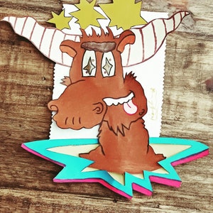 Puede incluir: Una ilustración de dibujos animados de un alce marrón con un cuerno rayado, que lleva una corona brillante y saca la lengua. El alce está de pie sobre una estrella azul, amarilla y rosa.