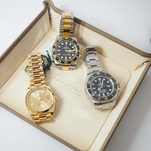 May include: Three luxury wristwatches in a brown leather watch tray. The watches have gold, silver, and black bands. The watch faces are black, gold, and silver. The tray has the words "Timepiece Temptations" printed on it.