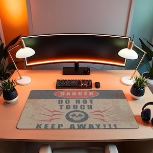 Könnte beinhalten: Ein Computertisch-Setup mit einem gebogenen Monitor, einer Tastatur und einer Maus. Ein rechteckiges Mauspad mit einem Used-Look-Design zeigt den Text "DANGER DO NOT TOUCH KEEP AWAY!!!" in Rot und Weiß mit einer Totenkopf-Grafik. Zwei Schreibtischlampen und Pflanzen flankieren den Monitor.