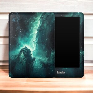 May include: A Kindle e-reader with a protective cover featuring a teal and black nebula design. The cover wraps around the device, displaying a cosmic scene with stars and swirling clouds. The Kindle logo is visible.