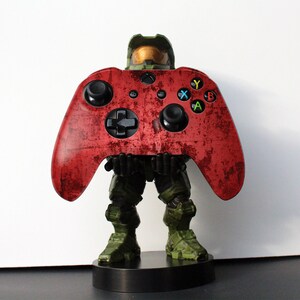 May include: A red Xbox controller with a distressed finish is held by a green and brown figure. The controller has black buttons and colored action buttons. The figure is standing on a black base. Gaming accessory.