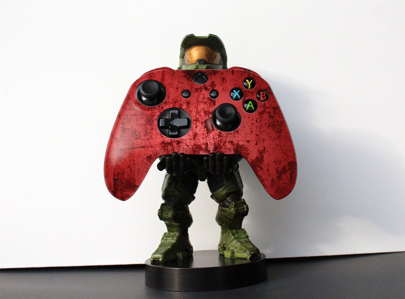 May include: A red Xbox controller with a distressed finish is held by a green and brown figure. The controller has black buttons and colored action buttons. The figure is standing on a black base. Gaming accessory.