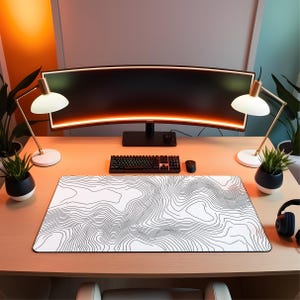 May include: A rectangular desk mat featuring a white surface with a black topographic map line pattern. The mat is positioned on a light wood desk in front of a curved computer monitor, a black mechanical keyboard, and a black computer mouse.