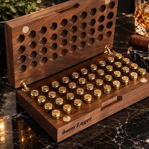 Puede incluir: Una caja de almacenamiento de madera marrón oscuro con tapa abatible, diseñada para contener 50 cartuchos de latón de 9mm Luger. La caja presenta una rejilla de cortes circulares en la tapa y la base, con el texto 9mm Luger grabado en el frente.
