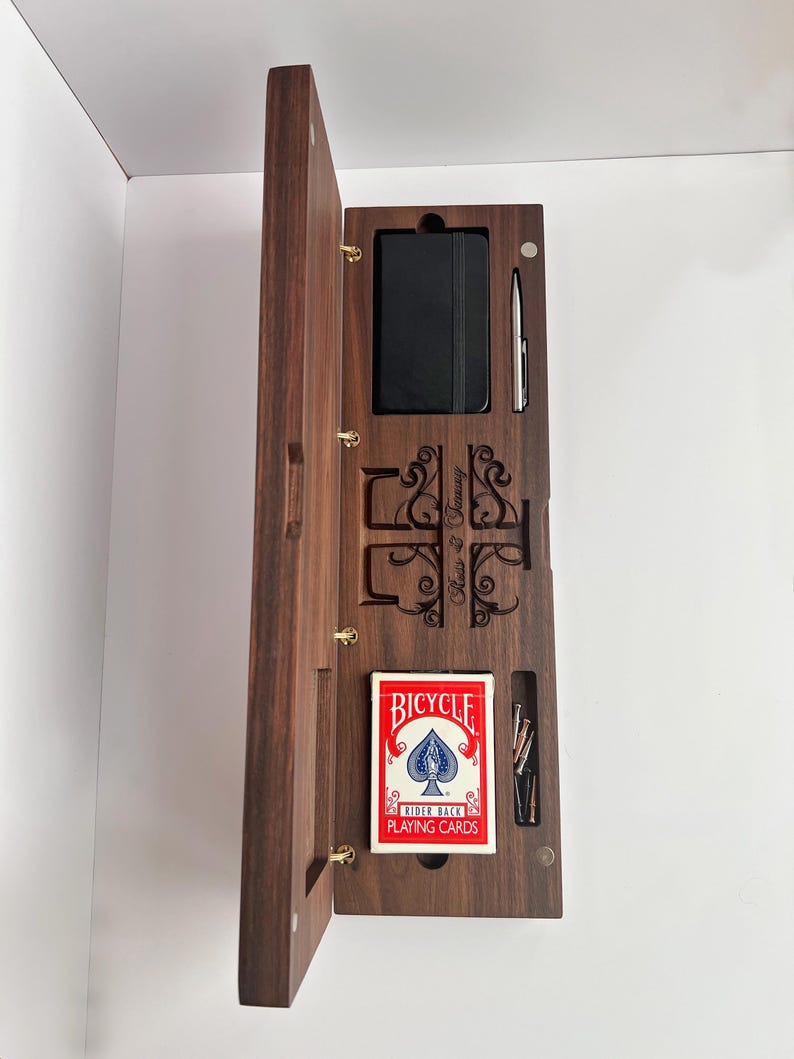 May include: A rectangular dark wood box with a hinged lid, containing a black notebook, a silver pen, a deck of Bicycle Rider Back playing cards, and several small metal pegs. The interior features laser-engraved scrollwork.