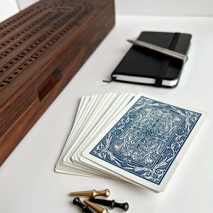 May include: A wooden cribbage board with a track, a deck of playing cards with blue and white ornate patterns, a small black notebook with a silver pen, and four metal cribbage pegs in gold and black finishes arranged on a surface.