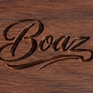 May include: A dark brown wooden surface featuring the word Boaz engraved in a cursive script font. The engraving has a darker, recessed appearance compared to the surrounding wood grain, which shows natural horizontal lines and texture.