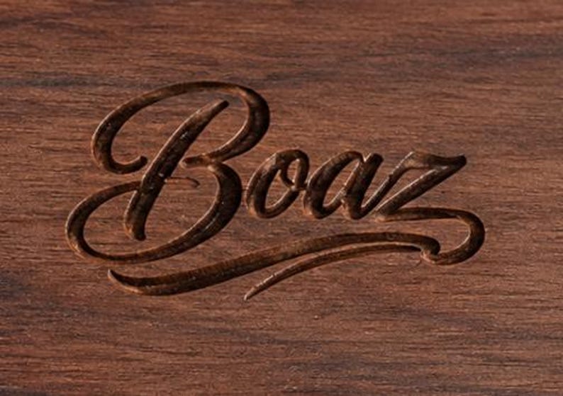 May include: A dark brown wooden surface featuring the word Boaz engraved in a cursive script font. The engraving has a darker, recessed appearance compared to the surrounding wood grain, which shows natural horizontal lines and texture.