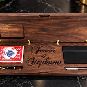 May include: A rectangular dark wood box with a hinged lid, featuring the engraved names Jenna & Stephano. Inside are a deck of red Bicycle playing cards, a black notebook with an elastic band, a silver pen, and several small metal pegs.