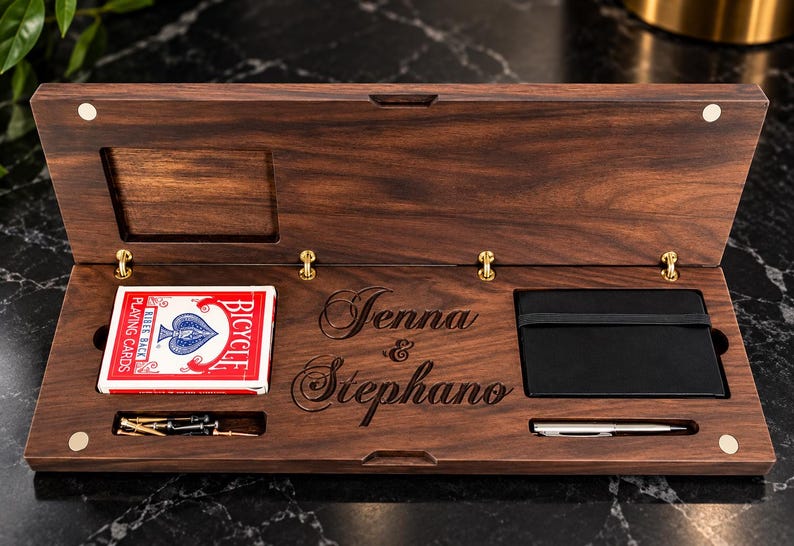 May include: A rectangular dark wood box with a hinged lid, featuring the engraved names Jenna & Stephano. Inside are a deck of red Bicycle playing cards, a black notebook with an elastic band, a silver pen, and several small metal pegs.