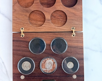 May include: A rectangular dark brown wooden coin display case with a hinged lid, featuring five circular recessed slots on the top half and five circular coin capsules on the bottom half, with one 1977 Eisenhower dollar coin visible.