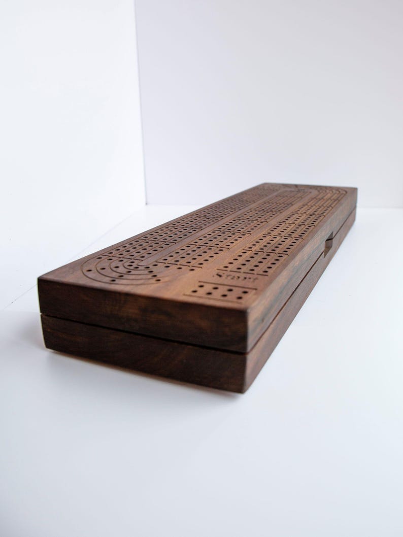 May include: A rectangular dark brown wooden cribbage board featuring a continuous track of drilled holes for scoring. The board has a smooth finish and includes the word Start engraved near the beginning of the game path on the top surface.