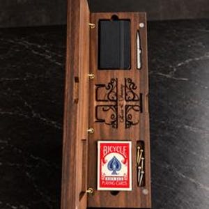 May include: A rectangular dark wood storage box with a hinged lid, containing a black notebook, a silver pen, a deck of red and white Bicycle playing cards, and several small metal pegs, featuring decorative laser-cut scrollwork.