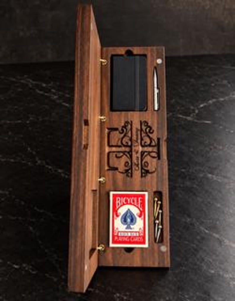May include: A rectangular dark wood storage box with a hinged lid, containing a black notebook, a silver pen, a deck of red and white Bicycle playing cards, and several small metal pegs, featuring decorative laser-cut scrollwork.