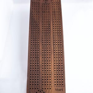 May include: A rectangular dark brown wooden cribbage board featuring three parallel tracks of holes for scoring. The board includes engraved text reading Start at the bottom and Out at the top, with a dedicated pegging area at the base.
