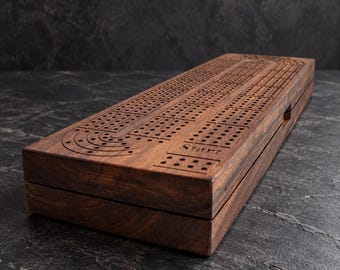 May include: A rectangular dark brown wooden cribbage board featuring a continuous track of drilled holes for scoring. The board has a smooth, polished finish with visible wood grain patterns and a clearly marked start section on the top surface.
