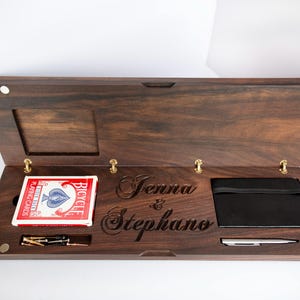 May include: A rectangular dark wood storage box with a hinged lid, featuring the engraved names Jenna & Stephano. Inside are a deck of red Bicycle playing cards, a black notebook with an elastic band, a silver pen, and small metal pegs.