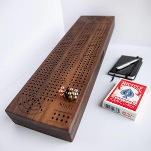 May include: A rectangular dark wood cribbage board with a grid of drilled holes and the word Start engraved on the surface. Five metal pegs in copper, black, gold, and silver are placed on the board. A deck of Bicycle playing cards and a black notebook with a silver pen are nearby.