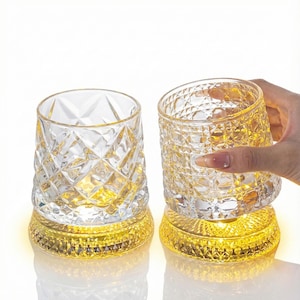 May include: Two clear, diamond-cut glass tumblers with a gold-colored base. One tumbler is being held by a hand. The bases emit a warm, yellow light, creating a decorative and elegant aesthetic. The glasses are likely for beverages.