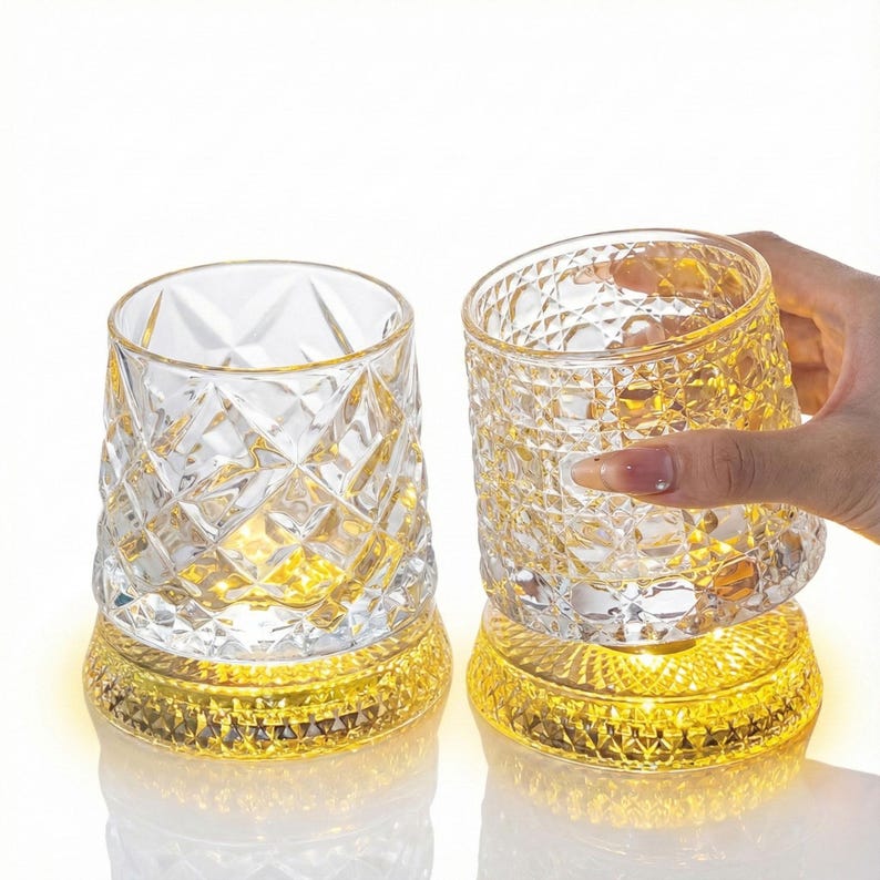 May include: Two clear, diamond-cut glass tumblers with a gold-colored base. One tumbler is being held by a hand. The bases emit a warm, yellow light, creating a decorative and elegant aesthetic. The glasses are likely for beverages.