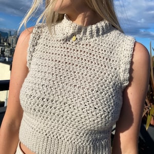 May include: A close-up of a woman wearing a sleeveless, crocheted, light gray crop top. The top has a high neckline and a button closure at the back of the neck.