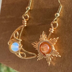 May include: Gold wire wrapped earrings with a crescent moon and sun design. The moon earring features a white opal stone and the sun earring features an orange carnelian stone.